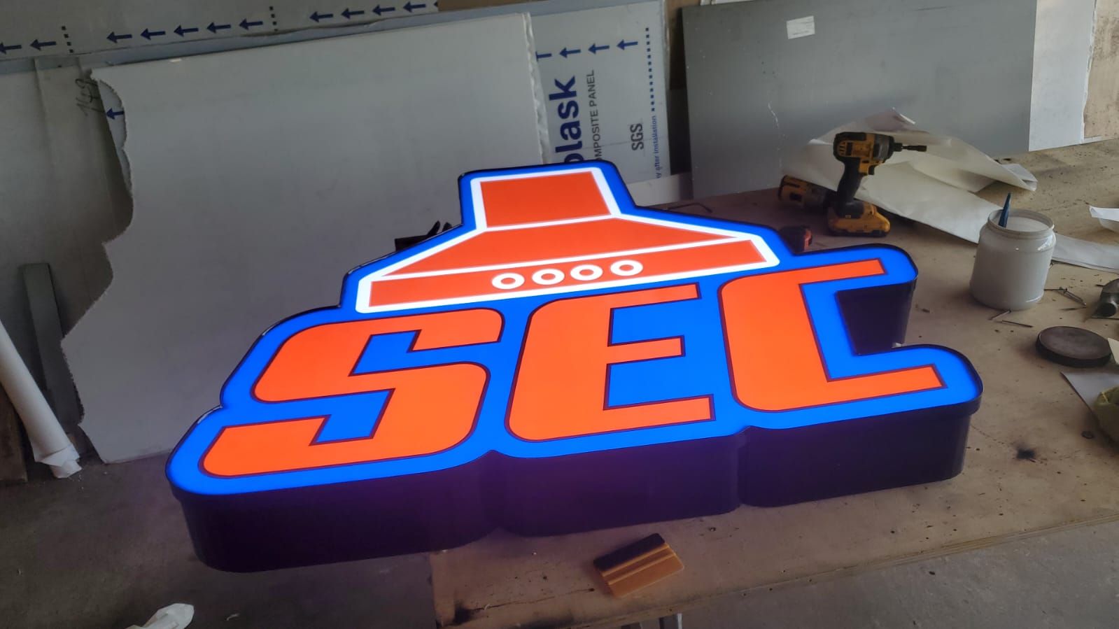 Charola luminosa recortada al contorno del logotipo SEC, encendida sobre el piso del taller junto a láminas de panel compuesto de aluminio.