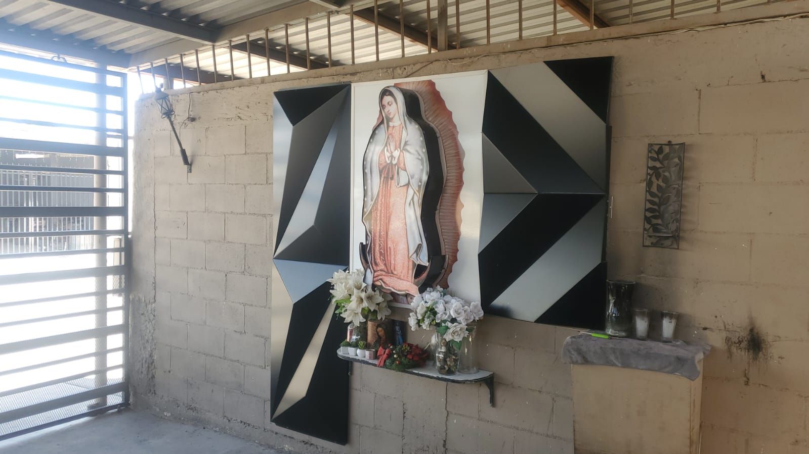 Retablo de la Virgen de Guadalupe enmarcado con paneles de aluminio facetados en negro y plata, montado sobre muro de block.