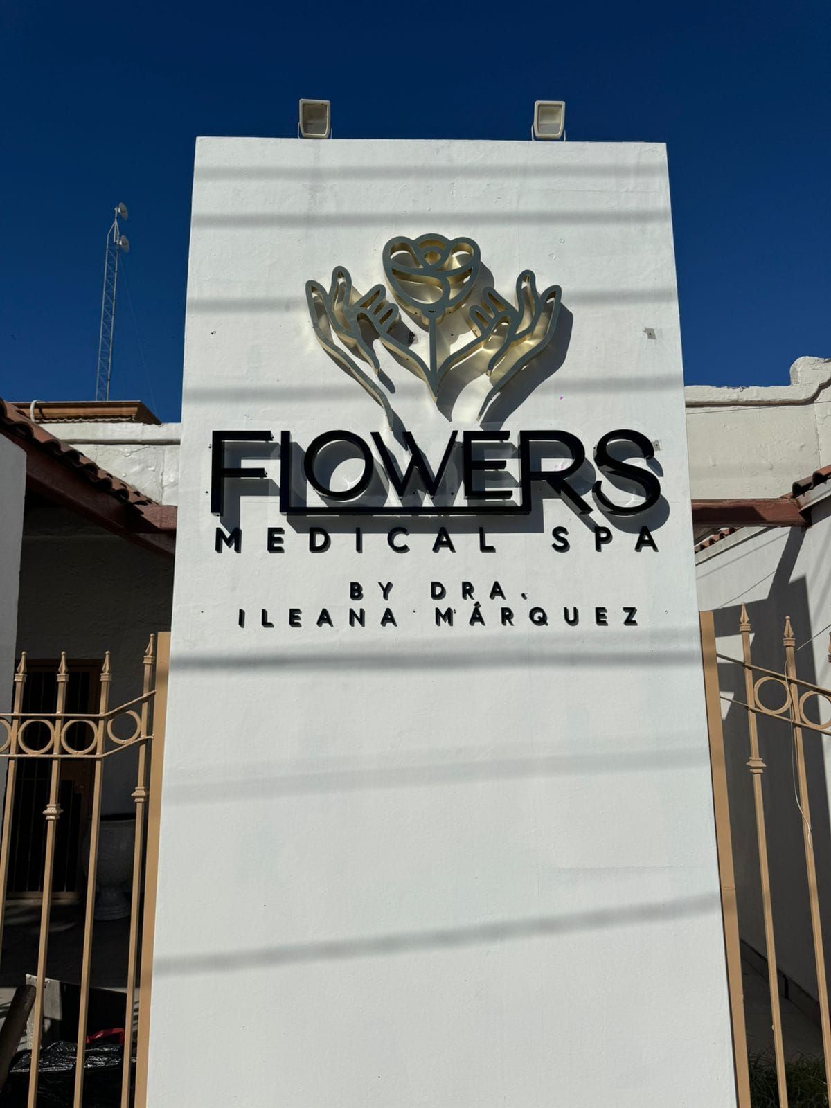 Tótem de Flowers Medical Spa: logotipo floral en acabado dorado con retroiluminación y letras negras de acrílico sobre muro blanco.