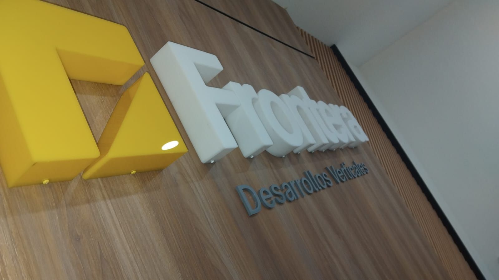 Letras corpóreas blancas y logotipo amarillo de Frontera Desarrollos Verticales sobre muro de listones de madera en interior.