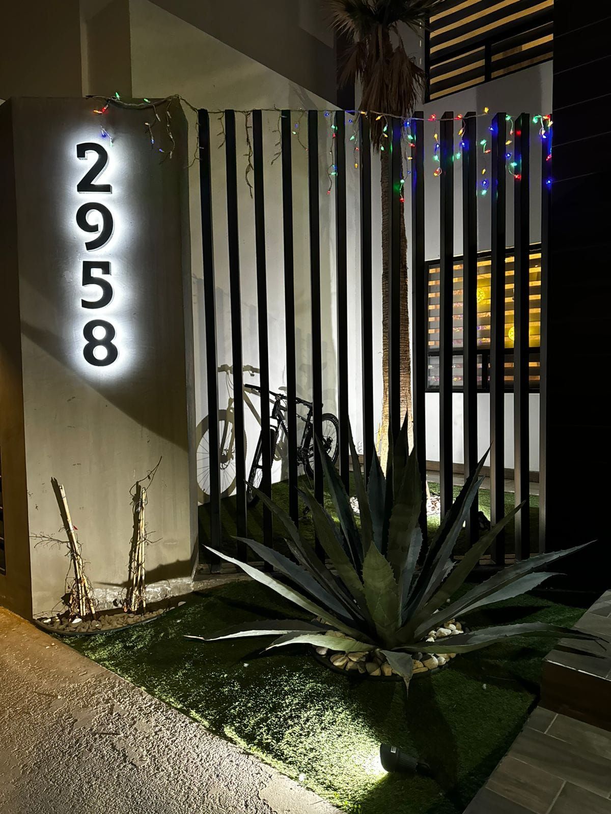 Número de casa 2958 en letras back-lit con halo blanco, montado en vertical sobre el muro de acceso de una residencia de noche.