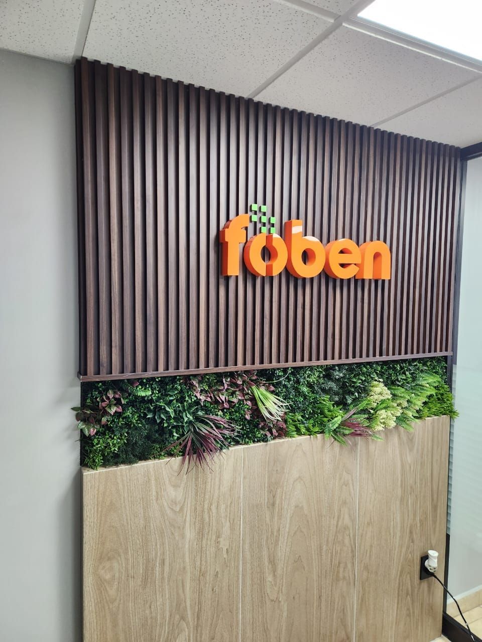 Logotipo corpóreo de fōben en naranja sobre panel de listones oscuros, con muro verde al pie.
