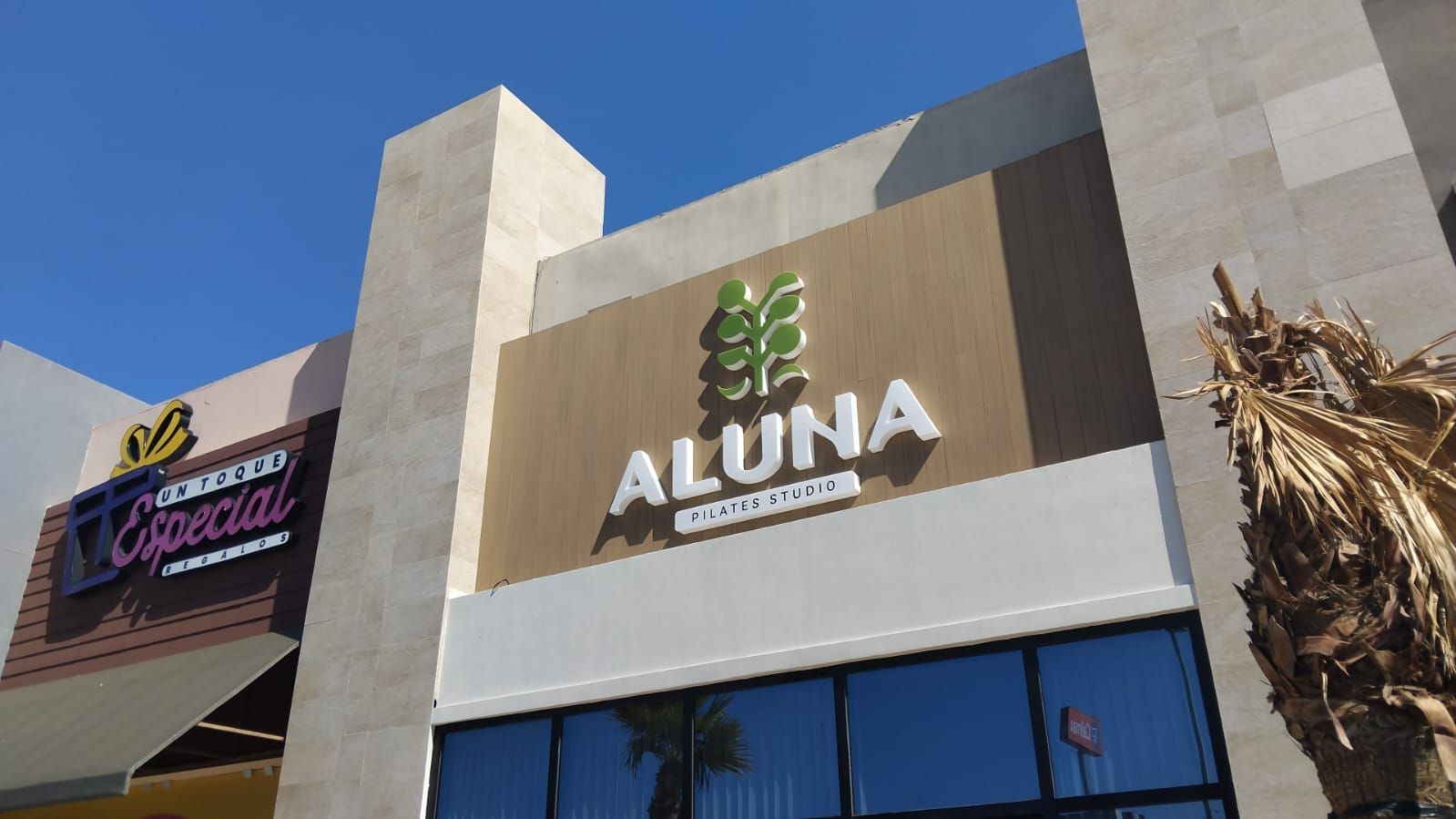 Letras corpóreas blancas y logotipo verde de Aluna Pilates Studio sobre panel de aluminio símil madera.