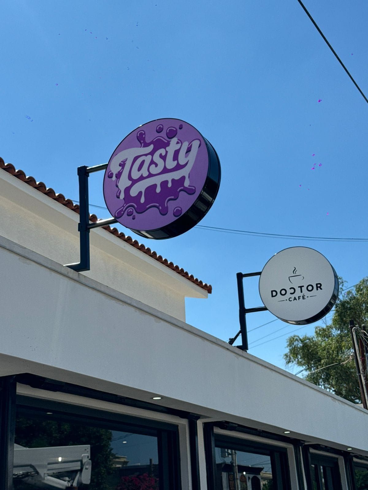 Dos banderas publicitarias circulares en voladizo, Tasty en morado y Doctor Café en blanco, sobre la cornisa del local.